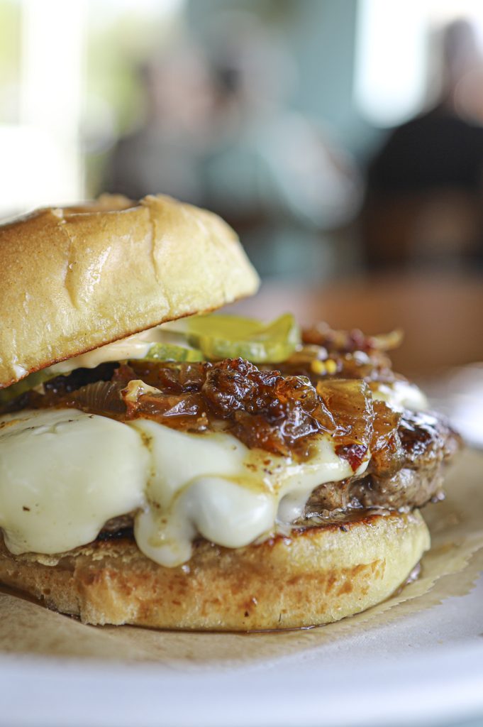 Close-Up of a Unique and Sumptuous gourmet burger topped with caramelized onions, pickled jalapenos, and melted Gruyère cheese, beautifully presented on a biodegradable plate, captured by YumSocial.co showcasing expert food photography in Fort Pierce, Florida and West Palm Beach, Florida